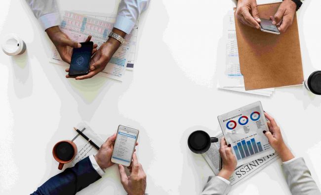 Vier Menschen sitzen am Tisch und schauen verschiedene Analysen an - auf dem Smartphone, in Mappen und auf Papier